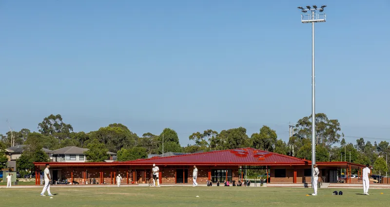 Long brick pavilion with red metal roof and deep eaves beside a lawn bowling green under clear sky