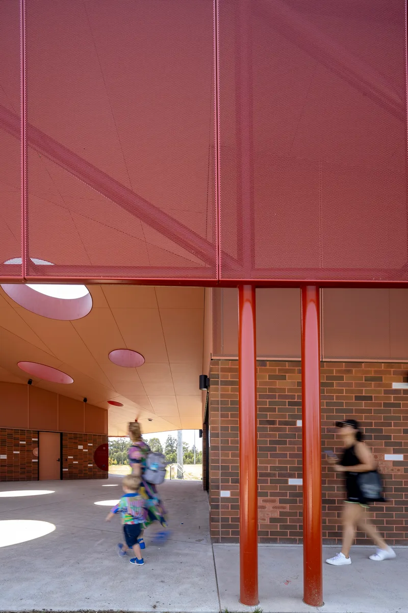 Covered breezeway with perforated red metal screen above and blurred figures walking through circular ceiling openings