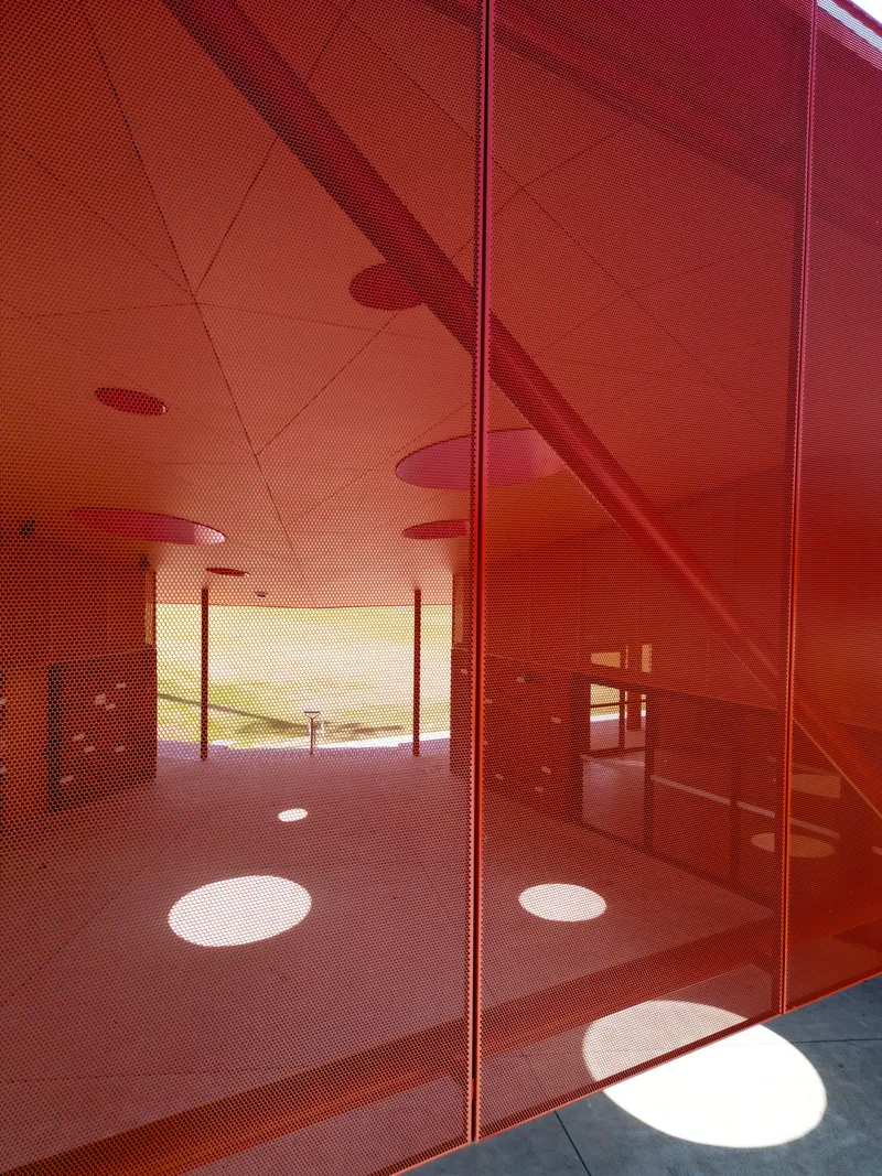 Reflections of circular ceiling apertures and columns on red perforated metal panels in afternoon sunlight