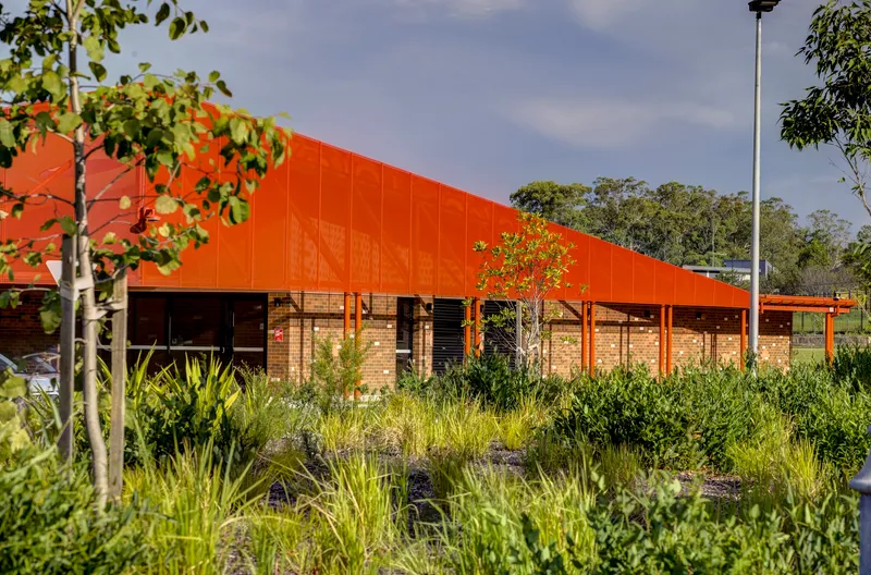 Long sloped orange metal canopy over brick walls viewed across native grasses and young trees