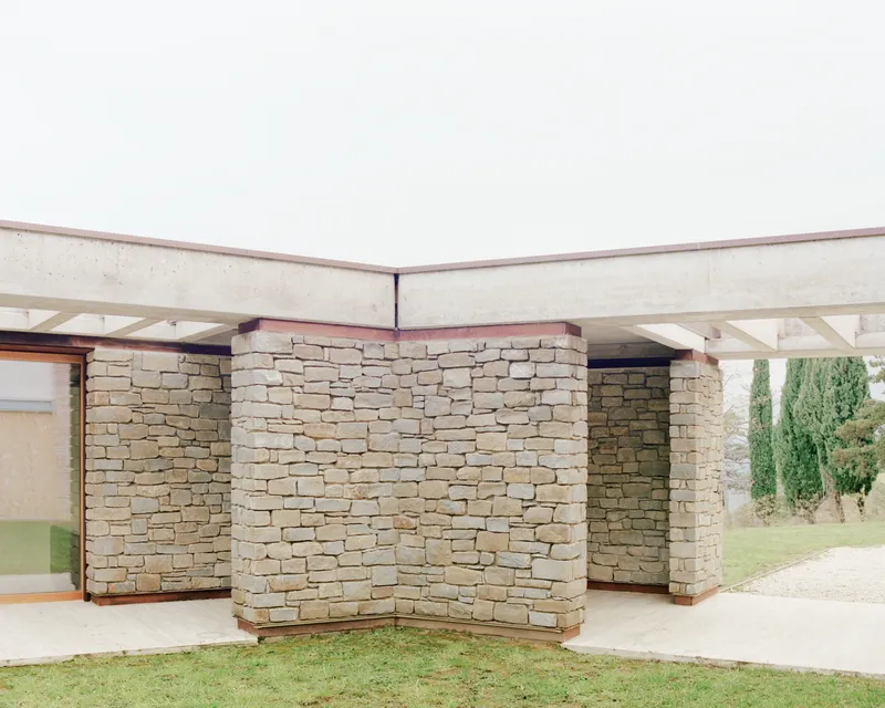 Layered stone walls beneath a flat concrete overhang with a lawn and cypress trees in the background