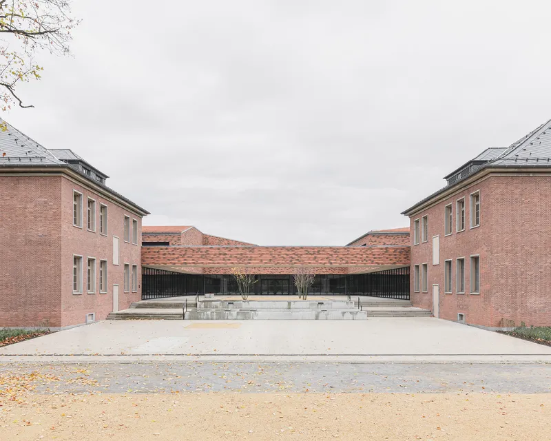 Symmetrical courtyard framed by pink brick volumes with a curved roof linking two wings under an overcast sky