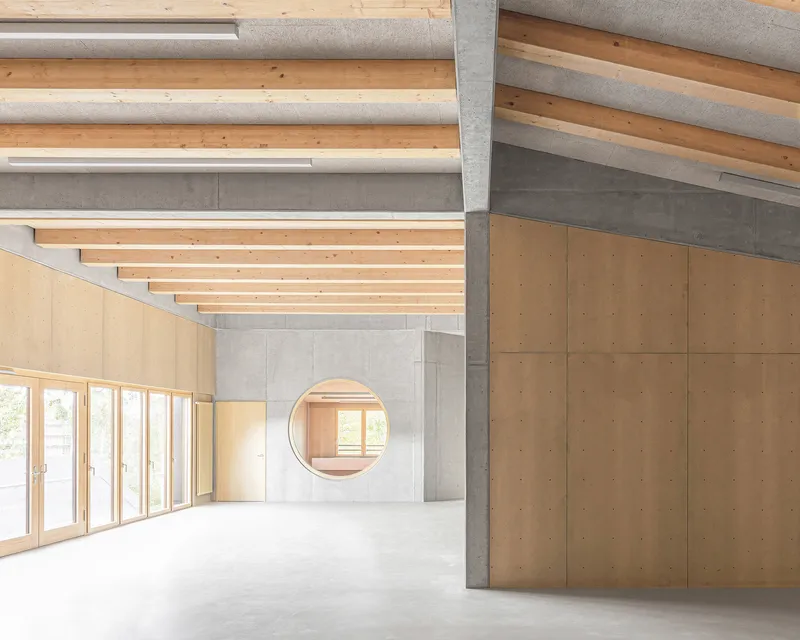 Interior space with exposed timber ceiling beams and a circular opening in a concrete wall framed by plywood panels