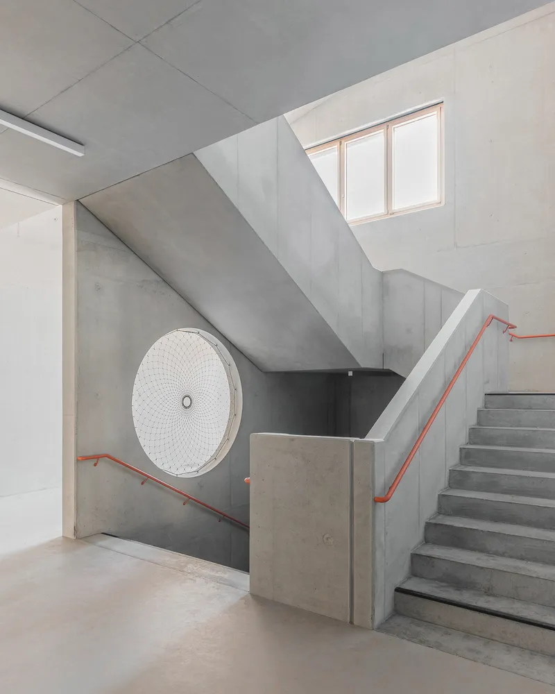 Concrete stairwell with orange handrails ascending toward a skylight above a circular decorative grille
