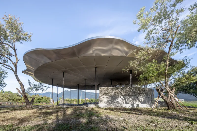 Undulating metal roof canopy supported by slender columns over a stone base surrounded by trees