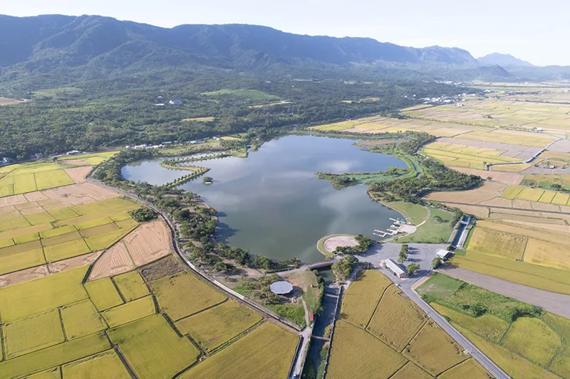 Aerial view of pavilions and curved roofs beside a reservoir surrounded by rice fields and mountains