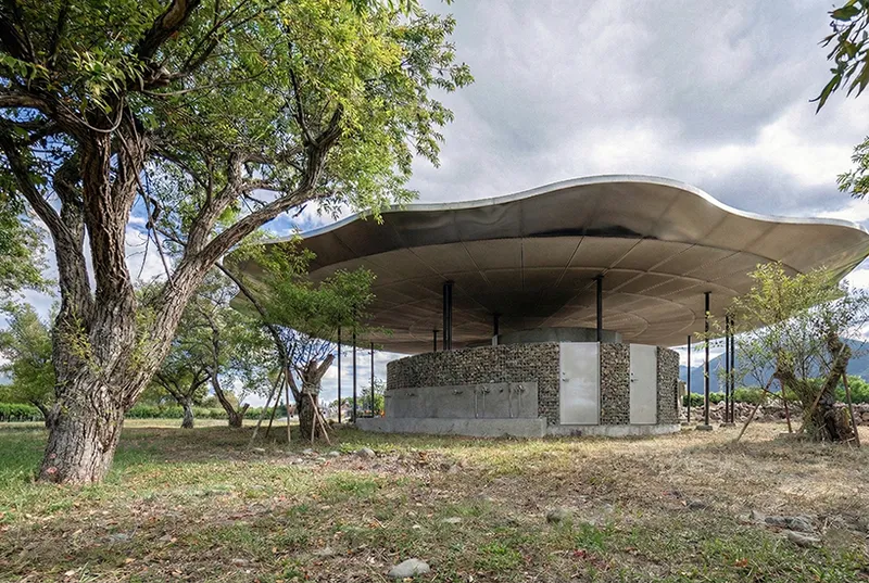 Pavilion with flowing metal canopy and stone-clad walls set among scattered trees under cloudy skies