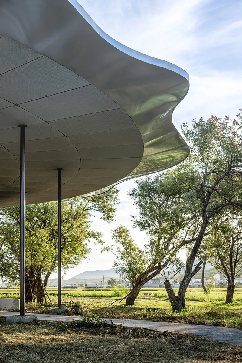 Underside view of the rippling metal roof edge supported by columns among mature trees at dusk