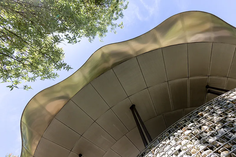 Upward view of the curved soffit panels and gabion wall beneath overhanging tree branches