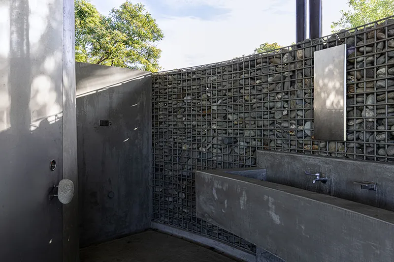 Outdoor shower enclosure with concrete walls and gabion basket screen casting leaf shadows