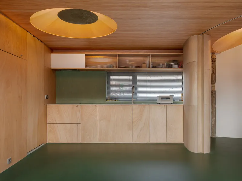 Plywood kitchenette with horizontal window and circular ceiling oculus above green floor