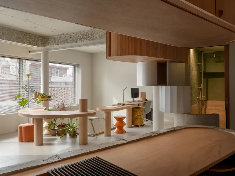 Living area with rounded table and curved wood desk platform overlooking a tiled bathroom in afternoon light