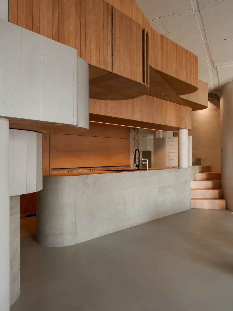Kitchen island with poured concrete base and timber staircase ascending beside suspended wood cabinetry