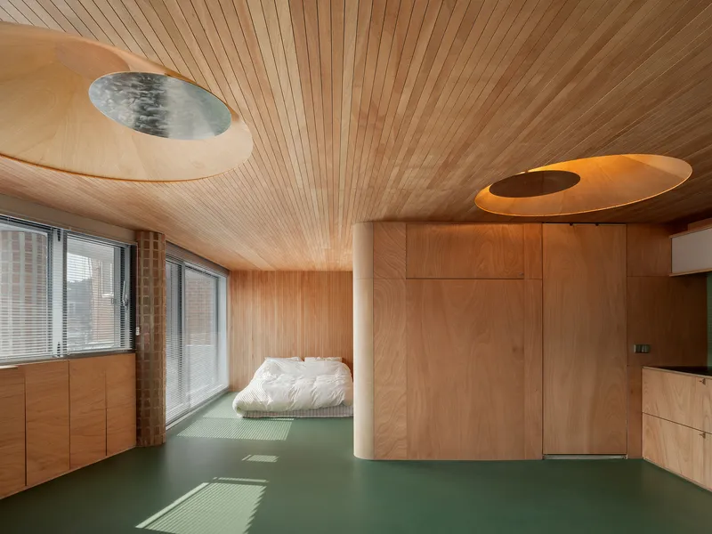 Bedroom with slatted timber ceiling, circular skylights, green floor, and white cylindrical column beside the low bed