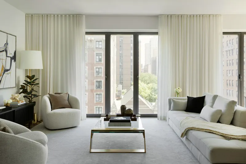 Living room with floor-to-ceiling windows, sheer curtains, and upholstered seating overlooking surrounding buildings