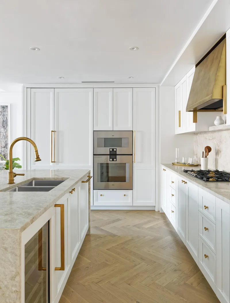 White paneled kitchen with brass fixtures and herringbone oak flooring under recessed lighting