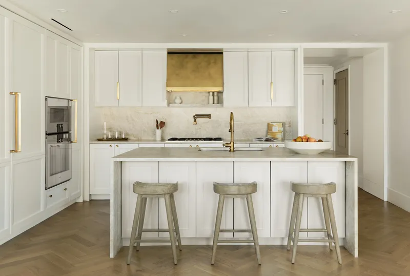 Kitchen island with three upholstered stools and brass range hood against white cabinetry