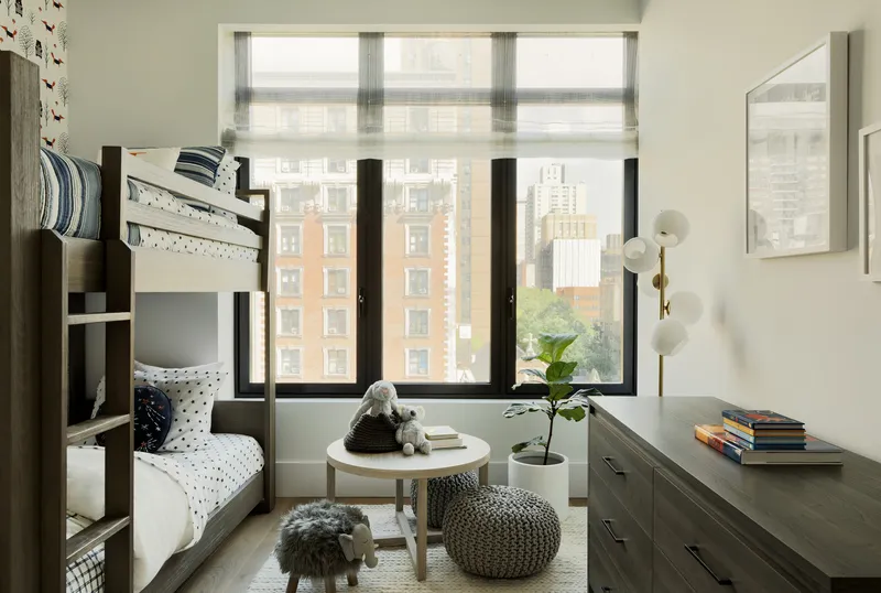 Bedroom with bunk bed and black-framed windows overlooking urban apartment buildings through sheer roller shades