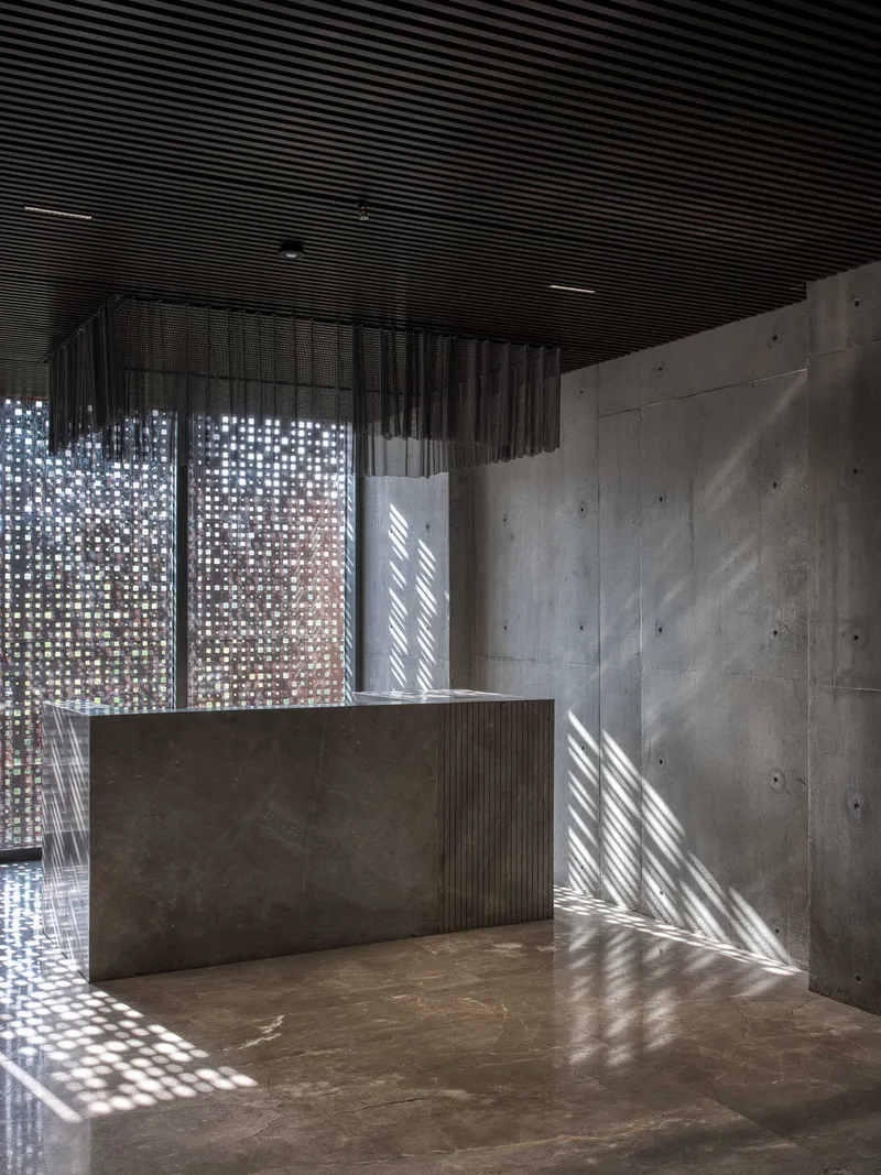 Reception desk with corrugated ribbed ceiling and perforated metal screen filtering dappled sunlight across concrete walls