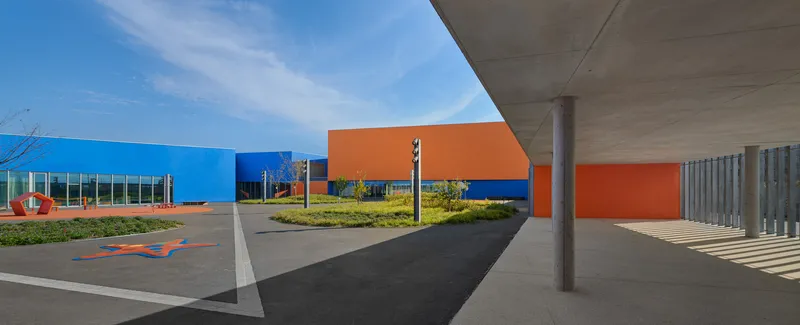 Covered entry drive beneath cantilevered concrete canopy with planted beds and colorful facade volumes beyond