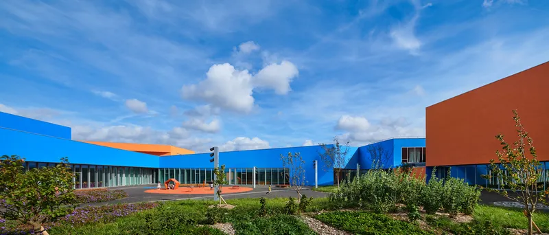 View across planted landscape toward the horizontal blue and orange volumes with glazed corridors in afternoon light