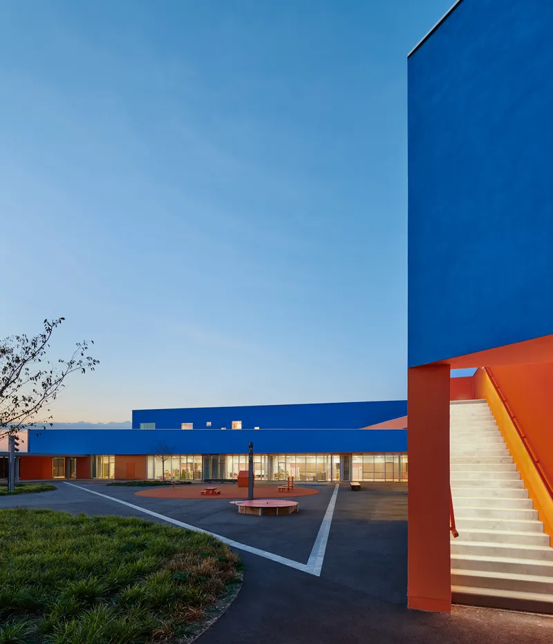 Exterior courtyard with orange staircase and benches at dusk with illuminated glazed corridors in background