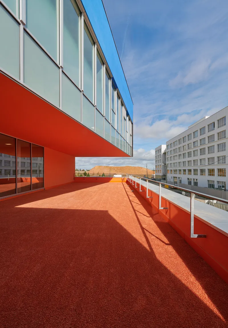 Orange rubberized rooftop terrace flanked by glazed corridor volume and white railing overlooking adjacent buildings