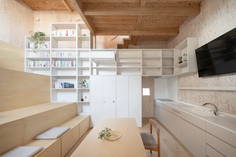 Living space with white storage wall, plywood stepped seating, and kitchen counter beneath timber ceiling