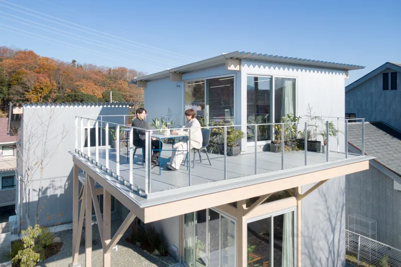 Elevated rooftop terrace with glass railing and corrugated metal siding, two figures seated in sunlight
