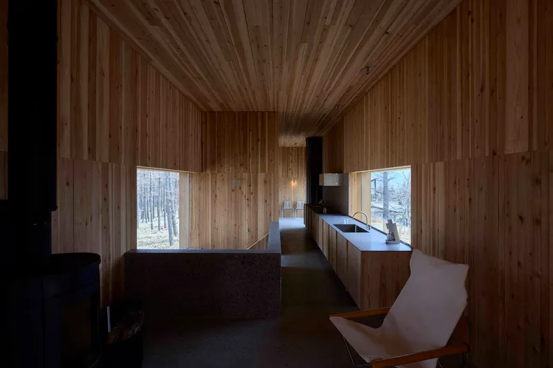 Kitchen and corridor with continuous timber surfaces and punched windows framing the winter forest