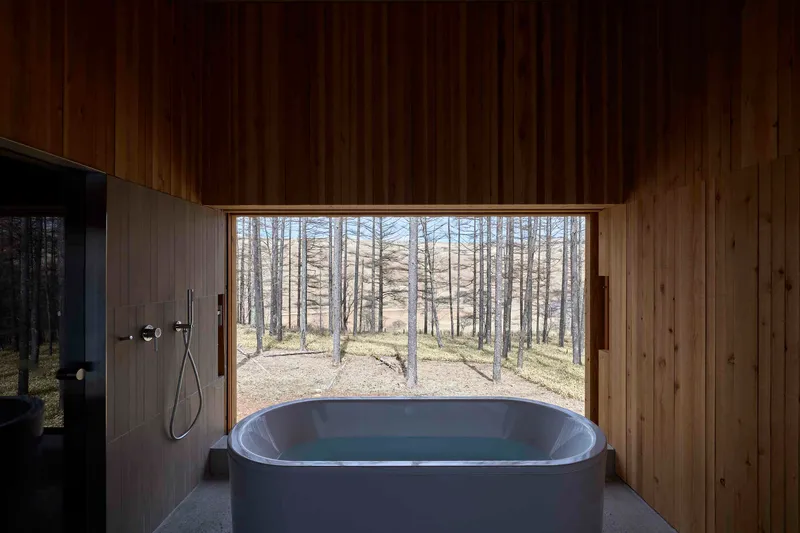 Freestanding bathtub beneath a horizontal window overlooking the sparse forest