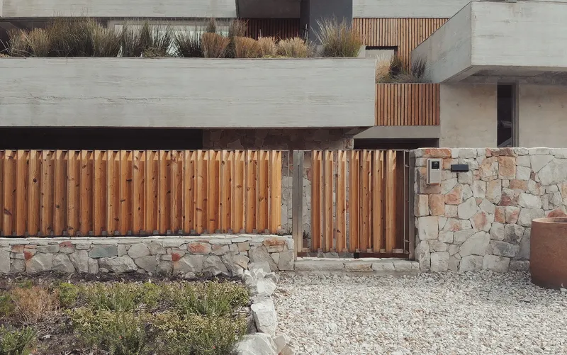 Entry courtyard with vertical timber slat gate and stone retaining walls flanking a gravel path
