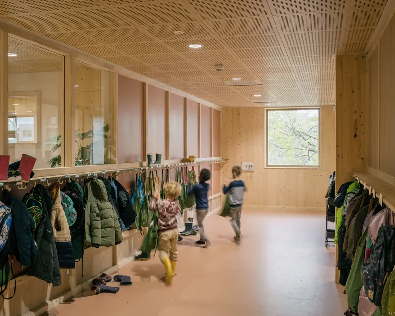 Corridor with perforated timber ceiling and children moving past coat hooks lining plywood walls