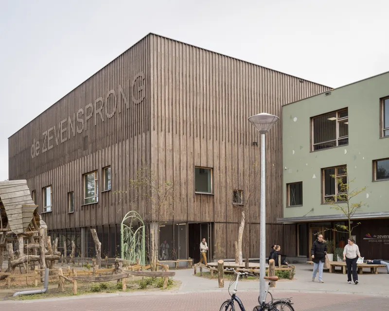 Vertical timber-clad facade with raised lettering adjoining a green plastered volume with a play area in front