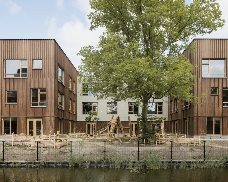 Courtyard view of timber-clad volumes flanking a white central block with a mature tree and wooden play structures