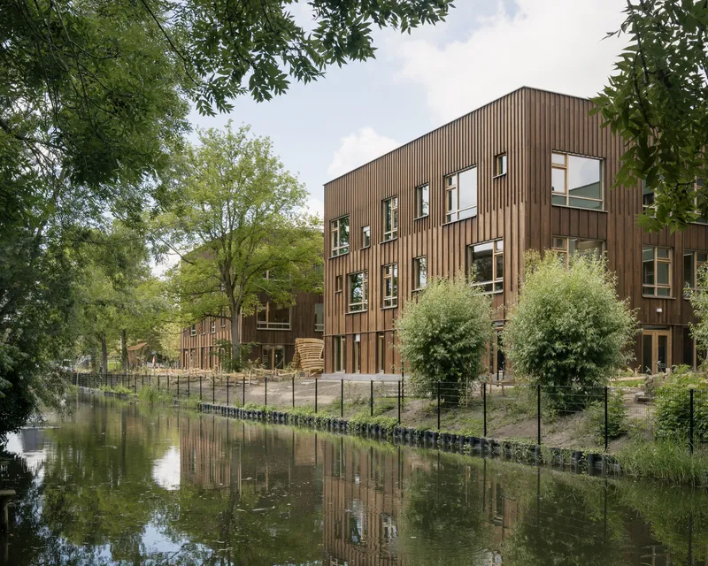 Vertical timber cladding facade with scattered window openings reflected in the adjacent canal