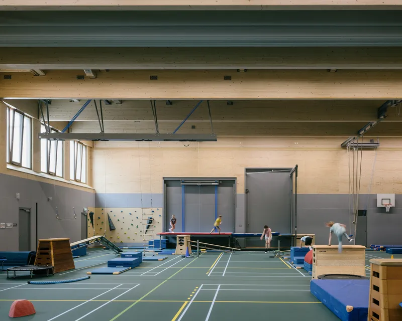 Interior gymnasium with exposed timber ceiling beams, plywood walls, and children playing among equipment