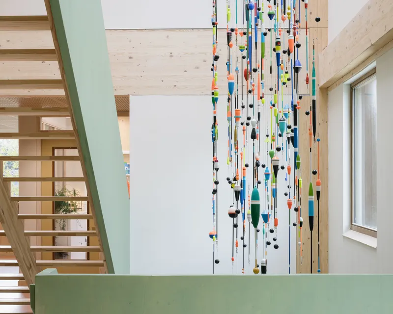 Double-height timber stairwell with painted green guardrail and hanging installation of colorful fishing floats