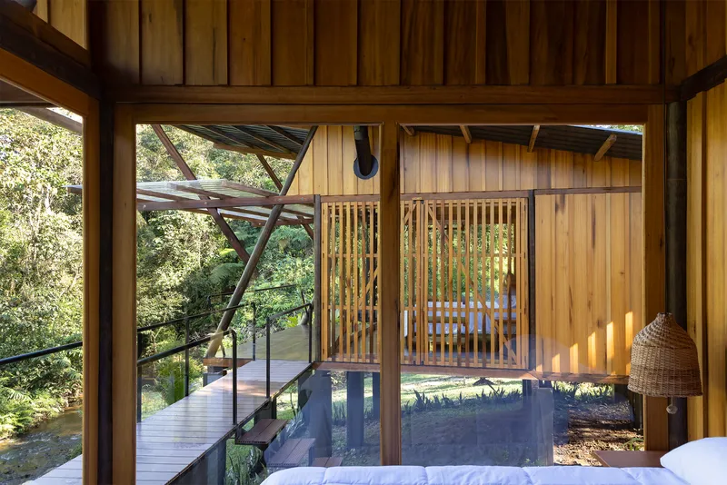 View from bedroom through timber-framed glazing to exterior walkway with diagonal bracing and forest beyond