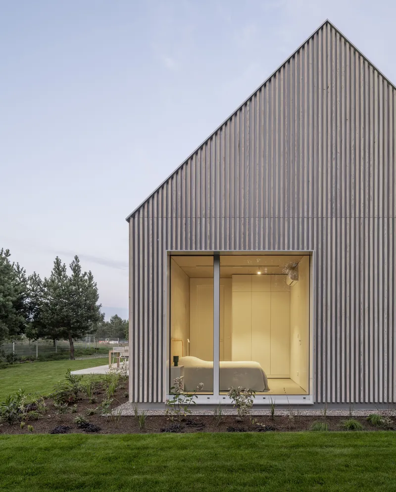 Gabled timber-clad volume with full-height glazing revealing illuminated bedroom at twilight