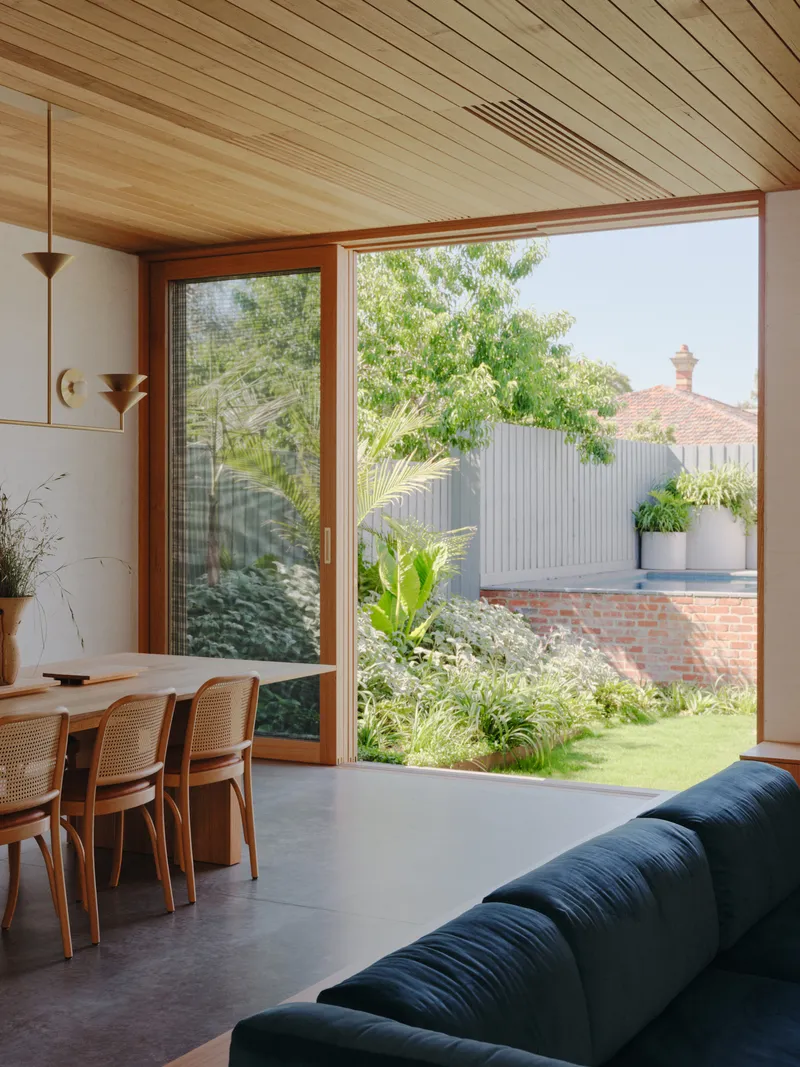 Dining area with corner glazing overlooking a planted garden with trees and corrugated metal fencing