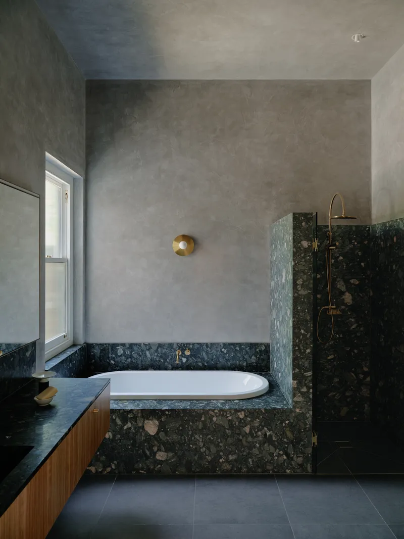 Bathroom with troweled grey plaster walls and dark terrazzo cladding the tub and shower enclosure