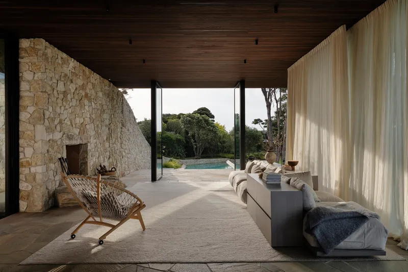 Open living area with full-height sheer curtains and irregular stone fireplace wall beneath a dark timber ceiling