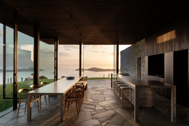 Dining and kitchen pavilion with irregular stone flooring opening to coastal views at sunset