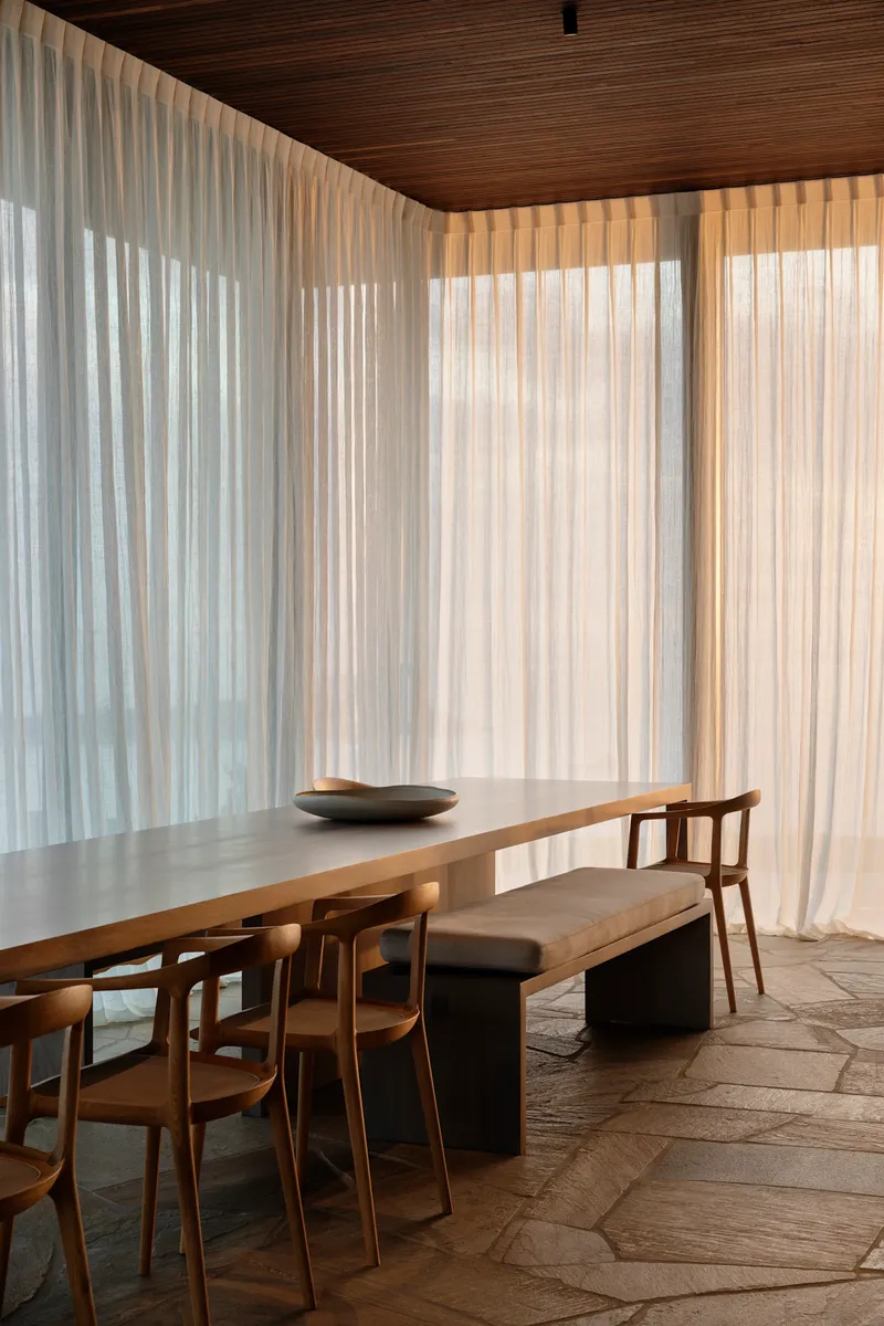 Dining room with timber table and irregular stone floor tiles beneath floor-to-ceiling sheer curtains
