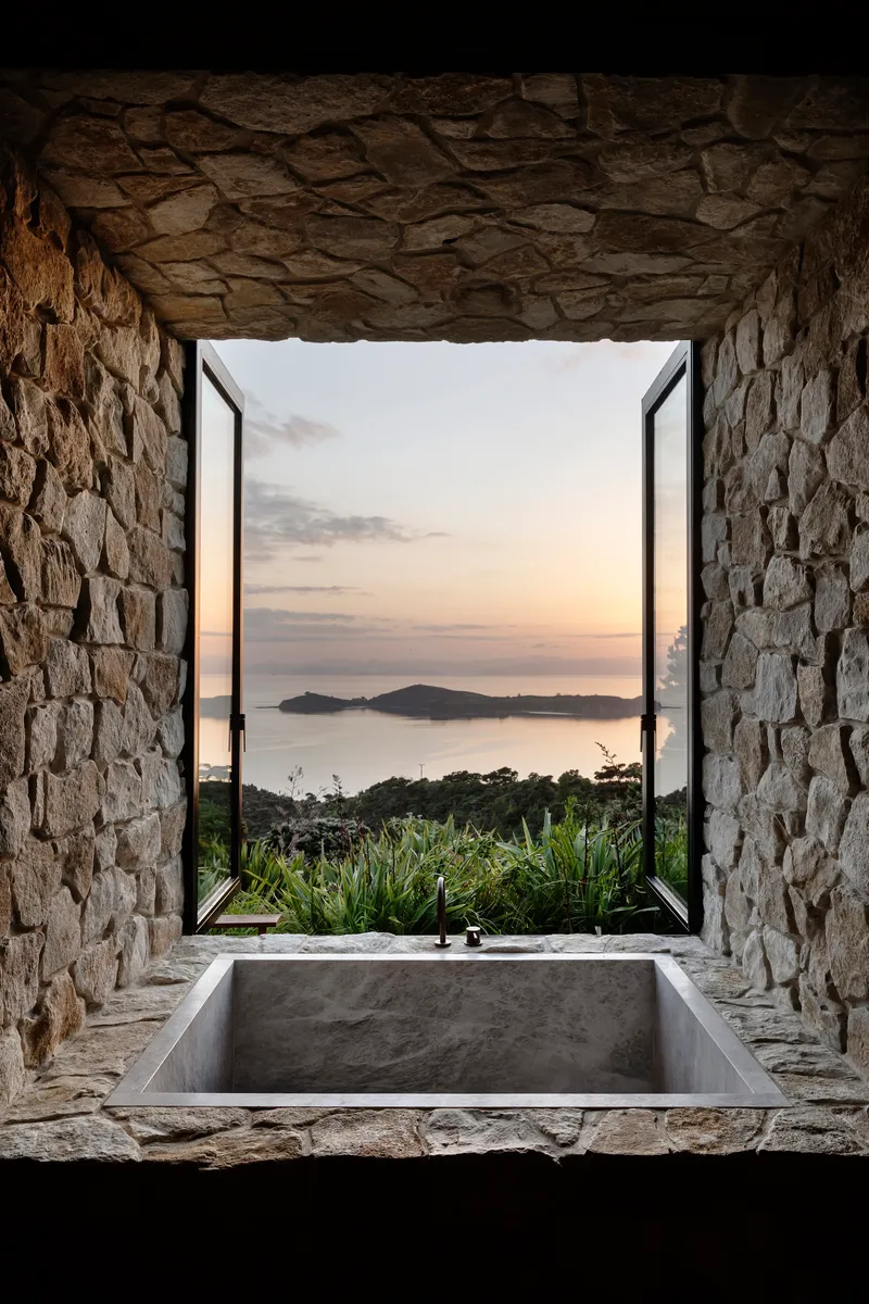 Stone-lined bathroom with sunken concrete tub framing sunset views over coastal water