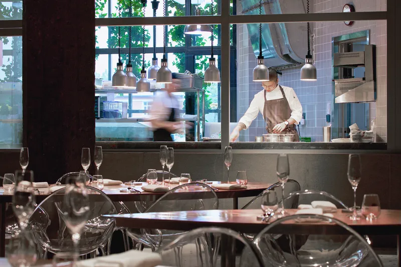 Restaurant dining area with clear acrylic chairs facing an open kitchen where a chef prepares food