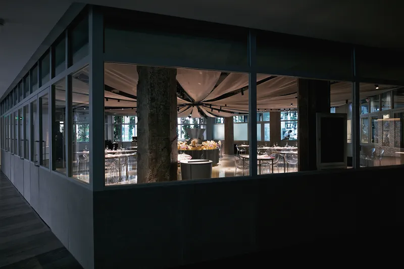 Exterior view through glazed facade showing the illuminated dining room with draped ceiling at dusk