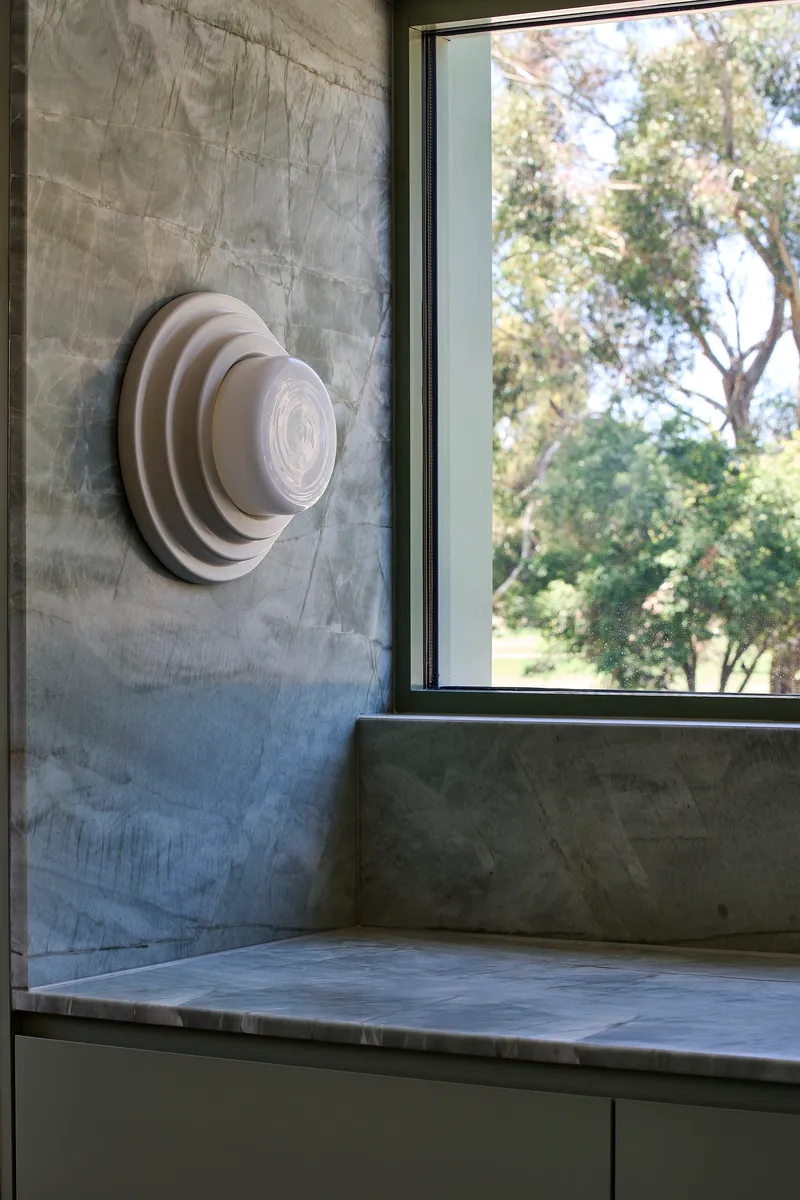 Circular wall-mounted light fixture on concrete wall beside a window framing leafy trees outside