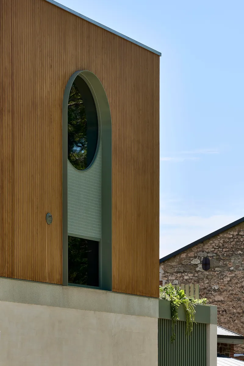 Vertical timber cladding facade with tall arched window opening and tiled green surround under blue sky